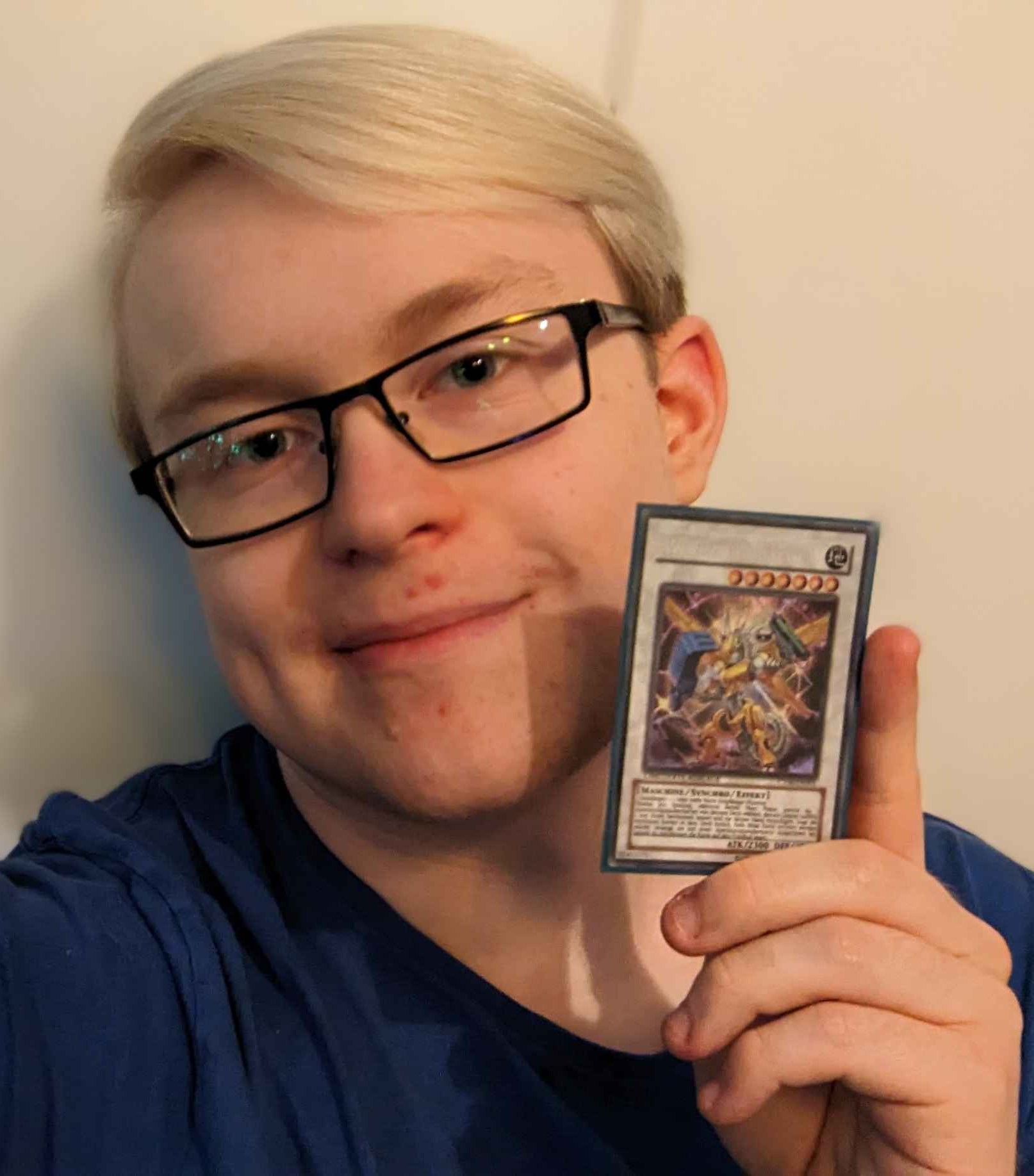 Ein Foto eines jungen Mannes mit Brille und weiß gefärbten Haaren.
Der junge Mann lächelt und hält eine Spielkarte hoch.